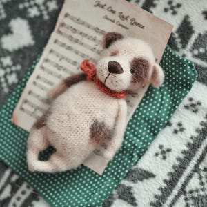 May include: A crocheted stuffed dog toy with brown spots and a red and orange scarf. The toy is lying on a green and white patterned fabric with sheet music in the background.