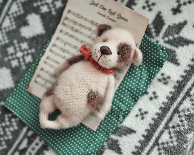 May include: A crocheted stuffed dog toy with brown spots and a red and orange scarf. The toy is lying on a green and white patterned fabric with sheet music in the background.