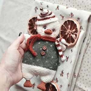 May include: A crocheted snowman toy with a red and white hat, a red scarf, and a candy cane in its hand. The snowman is wearing a green and white sweater and has a white face with black eyes and a black beak.