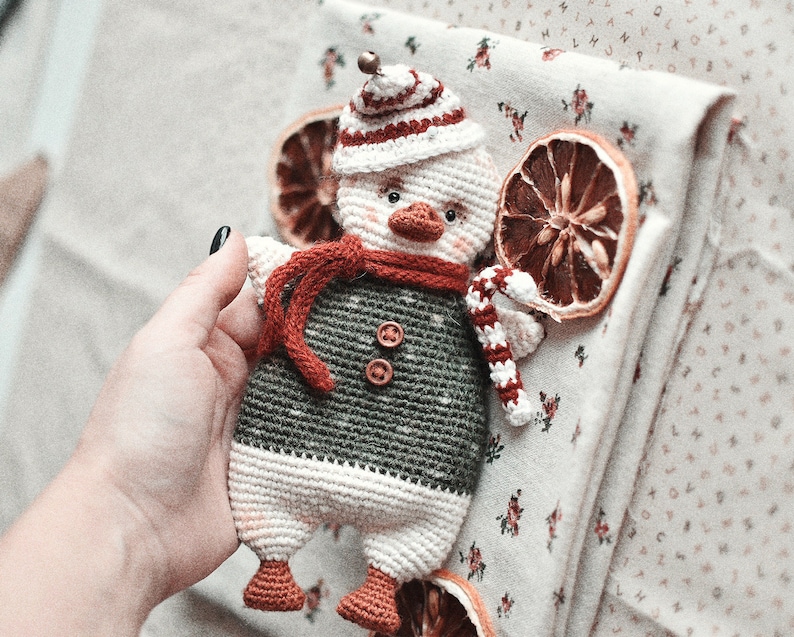 May include: A crocheted snowman toy with a red and white hat, a red scarf, and a candy cane in its hand. The snowman is wearing a green and white sweater and has a white face with black eyes and a black beak.