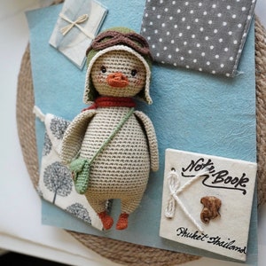 Pode incluir: Um pato de pel&uacute;cia de croch&ecirc; vestindo um chap&eacute;u de aviador verde e uma echarpe marrom. O pato est&aacute; segurando uma pequena bolsa verde e est&aacute; em p&eacute; sobre uma superf&iacute;cie azul. O fundo inclui um tecido de bolinhas brancas e cinzas, um tecido branco com um desenho de &aacute;rvore e uma placa branca com o texto "Note Book Phuket Thailand".