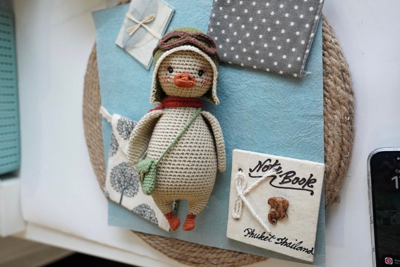 Pode incluir: Um pato de pel&uacute;cia de croch&ecirc; vestindo um chap&eacute;u de aviador verde e uma echarpe marrom. O pato est&aacute; segurando uma pequena bolsa verde e est&aacute; em p&eacute; sobre uma superf&iacute;cie azul. O fundo inclui um tecido de bolinhas brancas e cinzas, um tecido branco com um desenho de &aacute;rvore e uma placa branca com o texto "Note Book Phuket Thailand".