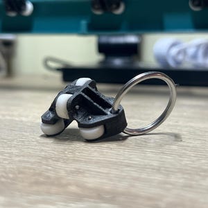 Puede incluir: Un componente de plástico negro con cuatro rodillos blancos y un anillo de metal plateado. El objeto está sobre una superficie de madera de color claro, con objetos borrosos en el fondo.
