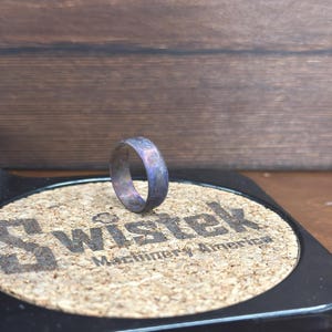 May include: A dark gray metal ring with a purple iridescent finish. The ring is sitting on a cork coaster with the text "Wisstek Machinery America".