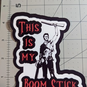 Könnte beinhalten: Ein weißer Aufkleber mit schwarzem Rand zeigt eine Silhouette einer Person, die eine Kettensäge und ein Gewehr hält. Der Text "This is my Boom Stick" ist in Rot geschrieben.