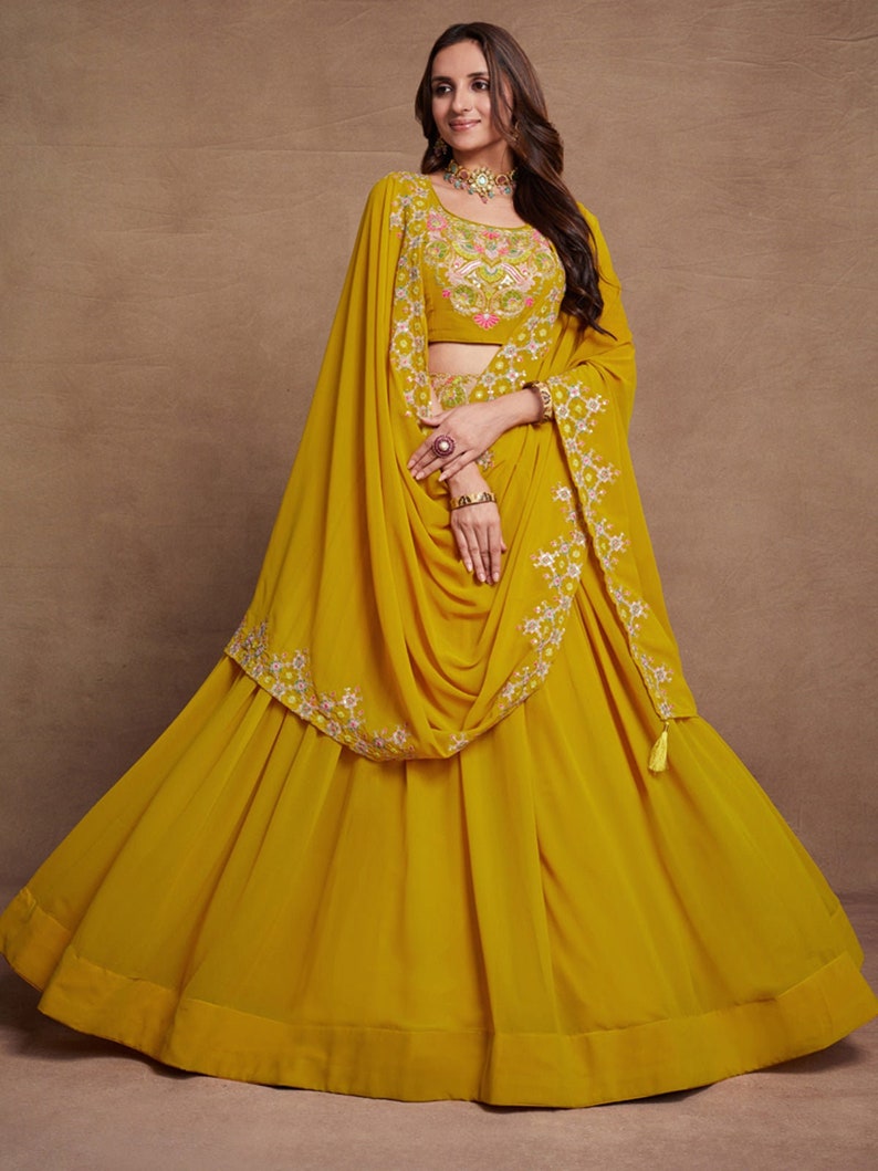 May include: A mustard yellow skirt with a matching dupatta and a floral embroidered blouse. The skirt has a flared silhouette and a ruffled hem. The dupatta is draped over the shoulders and has a floral embroidered border. The blouse has a round neckline and short sleeves.