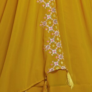 May include: A mustard yellow skirt with a geometric pattern embroidered on the side. The embroidery is in shades of pink, green, and white. The skirt has a tassel detail at the hem.