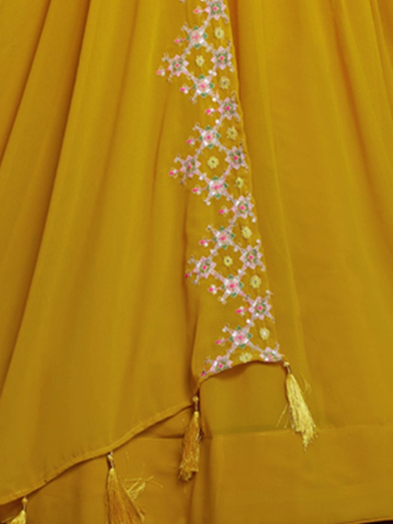 May include: A mustard yellow skirt with a geometric pattern embroidered on the side. The embroidery is in shades of pink, green, and white. The skirt has a tassel detail at the hem.