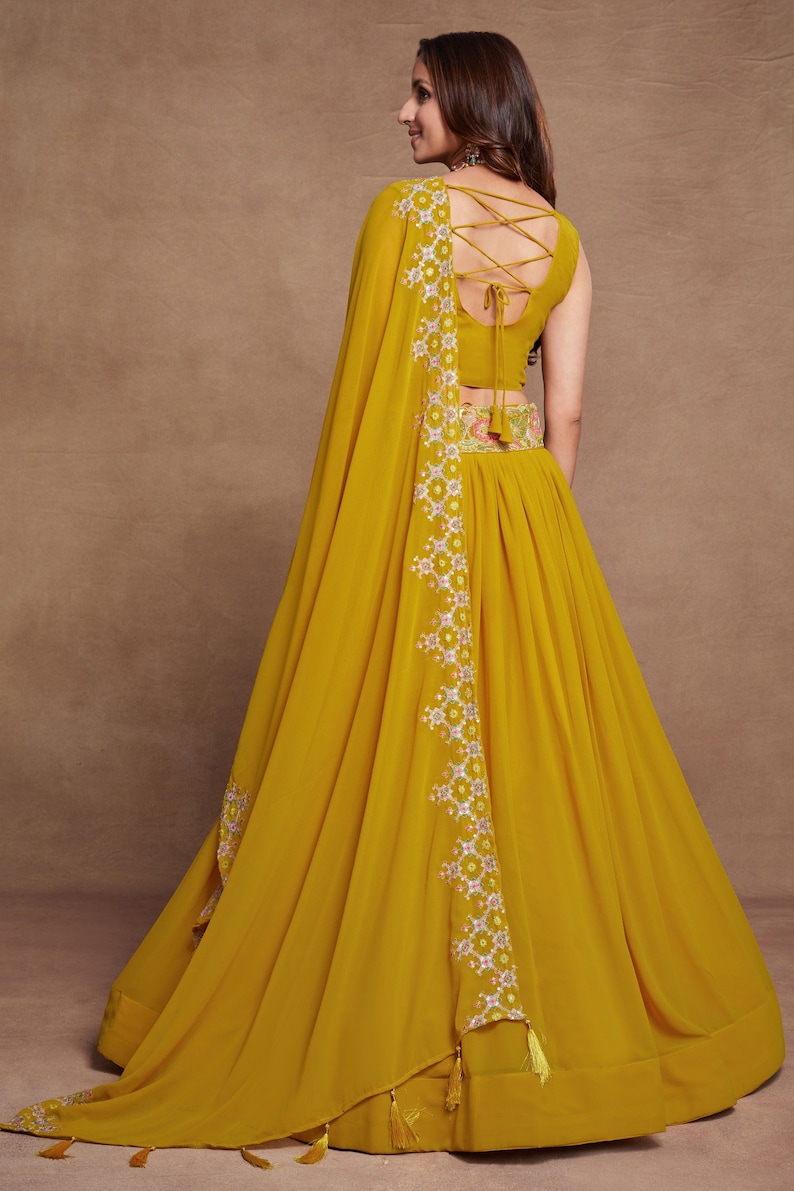 May include: A mustard yellow maxi skirt with a floral embroidered waistband and a matching yellow dupatta with a white and pink floral embroidered border. The dupatta has tassels on the edges.
