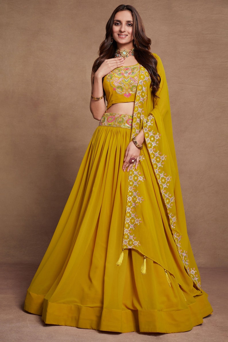 May include: A mustard yellow lehenga with floral embroidery and a matching crop top. The outfit includes a sheer dupatta with floral embroidery and tassels.