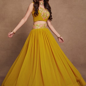 May include: A mustard yellow sleeveless crop top with floral embroidery and a matching mustard yellow skirt with pleats. The skirt has a gold and silver embroidered waistband.