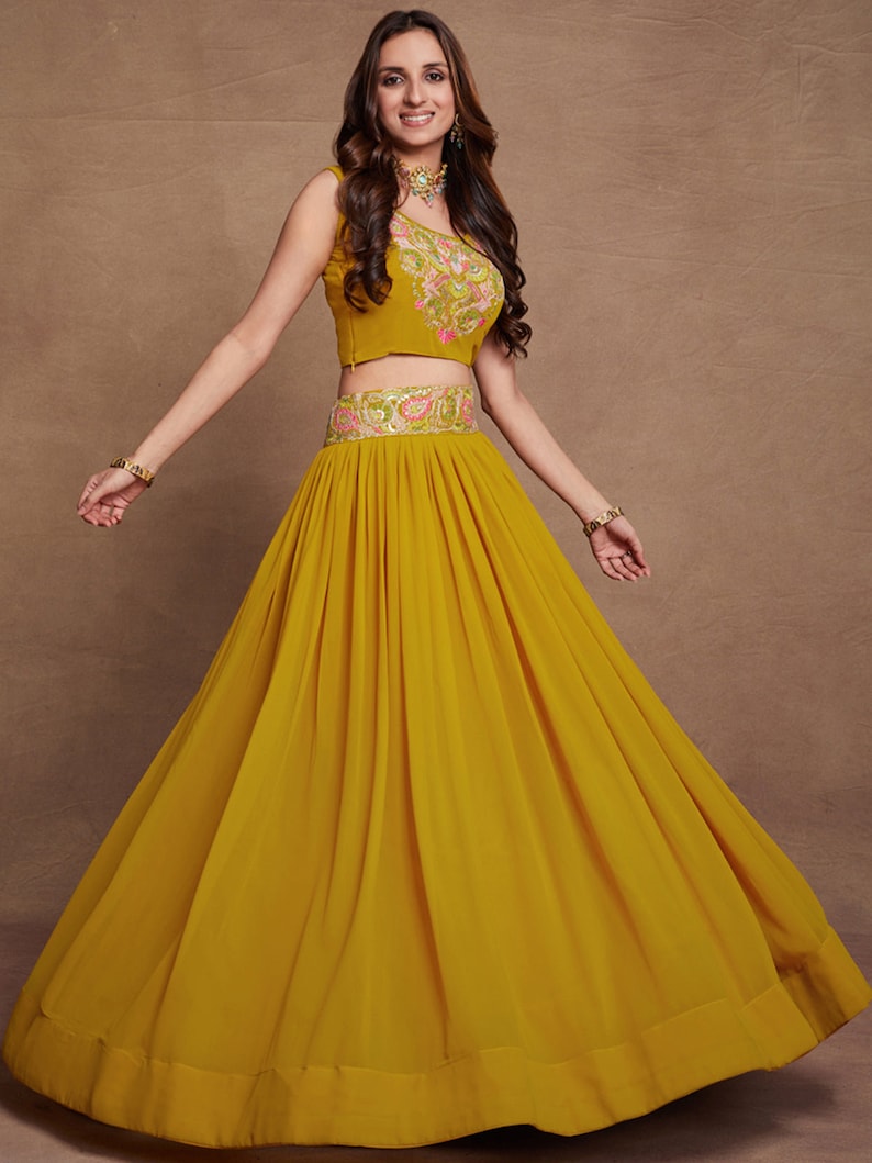 May include: A mustard yellow sleeveless crop top with floral embroidery and a matching mustard yellow skirt with pleats. The skirt has a gold and silver embroidered waistband.