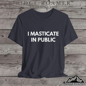 May include: Dark grey t-shirt with the white text "I Masticate In Public". The shirt is from Ridge Roamer Clothing Co. and is displayed against a wooden backdrop.