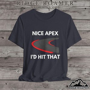 May include: Dark gray t-shirt with the text "NICE APEX I'D HIT THAT" above a graphic of a curved road with a red accent. The shirt has a crew neck and short sleeves. The Ridge Roamer Clothing Co. logo is at the top and bottom.