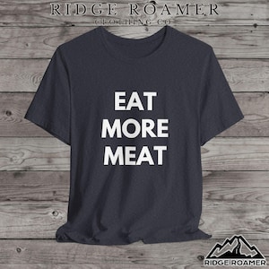 May include: Dark grey t-shirt with the white text "EAT MORE MEAT". The shirt is from Ridge Roamer Clothing Co. and is displayed against a wooden background.
