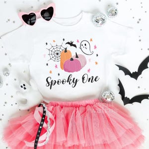 May include: White t-shirt with a pink and orange pumpkin design, a ghost, and the text "Spooky One". The shirt is paired with a pink tulle skirt.