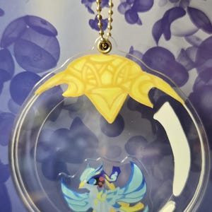 Könnte beinhalten: Ein transparenter Acryl-Schlüsselanhänger mit einer goldenen Kette. Im Inneren des Schlüsselanhängers befindet sich ein blauer und gelber Vogel vor weißem Hintergrund. Der Vogel hat einen gelben Schnabel und gelbe Füße.