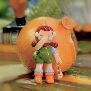 May include: A whimsical figurine of a person in green overalls, orange boots, and a red helmet, holding a small bag and a charm. The charm is a small egg with a chicken design. The figurine is set against a blurred background with an onion.
