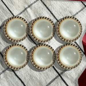 May include: Six white pearl-like buttons with a gold metal rim and a rope-like design. The buttons are arranged in two rows of three.