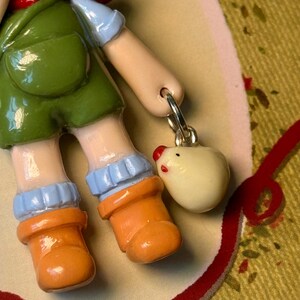 May include: A ceramic figurine of a child wearing green overalls, orange boots, and blue sleeves. A small, white egg-shaped charm with a red beak and eyes hangs from a silver ring.