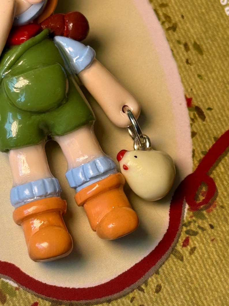 May include: A ceramic figurine of a child wearing green overalls, orange boots, and blue sleeves. A small, white egg-shaped charm with a red beak and eyes hangs from a silver ring.