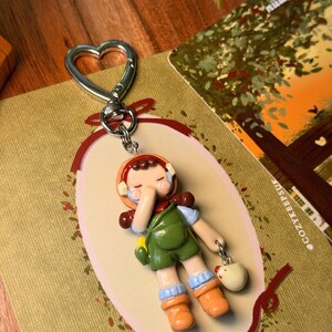 May include: A whimsical keychain featuring a miniature figurine of a child in a green jumpsuit, orange boots, and a brown hat. The keychain has a heart-shaped clasp and a small charm of a white chicken. The figurine is set against a decorative background.