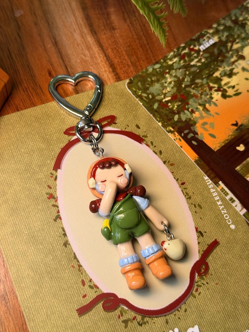 May include: A whimsical keychain featuring a miniature figurine of a child in a green jumpsuit, orange boots, and a brown hat. The keychain has a heart-shaped clasp and a small charm of a white chicken. The figurine is set against a decorative background.