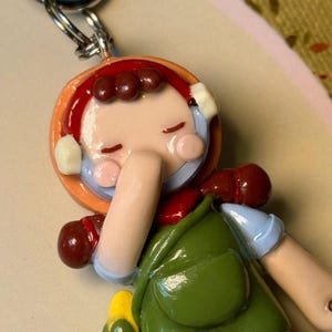 May include: A handmade keychain featuring a miniature figurine of a person in a green dress and red hair. The figurine has a cheerful expression and is attached to a silver-colored metal ring.