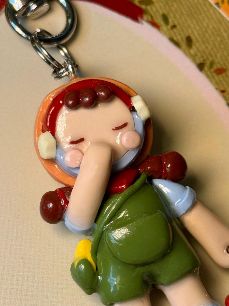 May include: A handmade keychain featuring a miniature figurine of a person in a green dress and red hair. The figurine has a cheerful expression and is attached to a silver-colored metal ring.