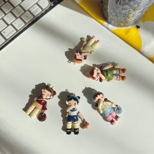 May include: A collection of six small, colorful doll figurines. The dolls are in various poses and outfits, including a maroon jacket, a blue and white uniform, and a green dress. Each doll has a small loop for hanging.