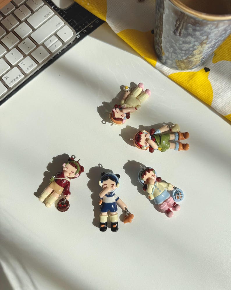 May include: A collection of six small, colorful doll figurines. The dolls are in various poses and outfits, including a maroon jacket, a blue and white uniform, and a green dress. Each doll has a small loop for hanging.