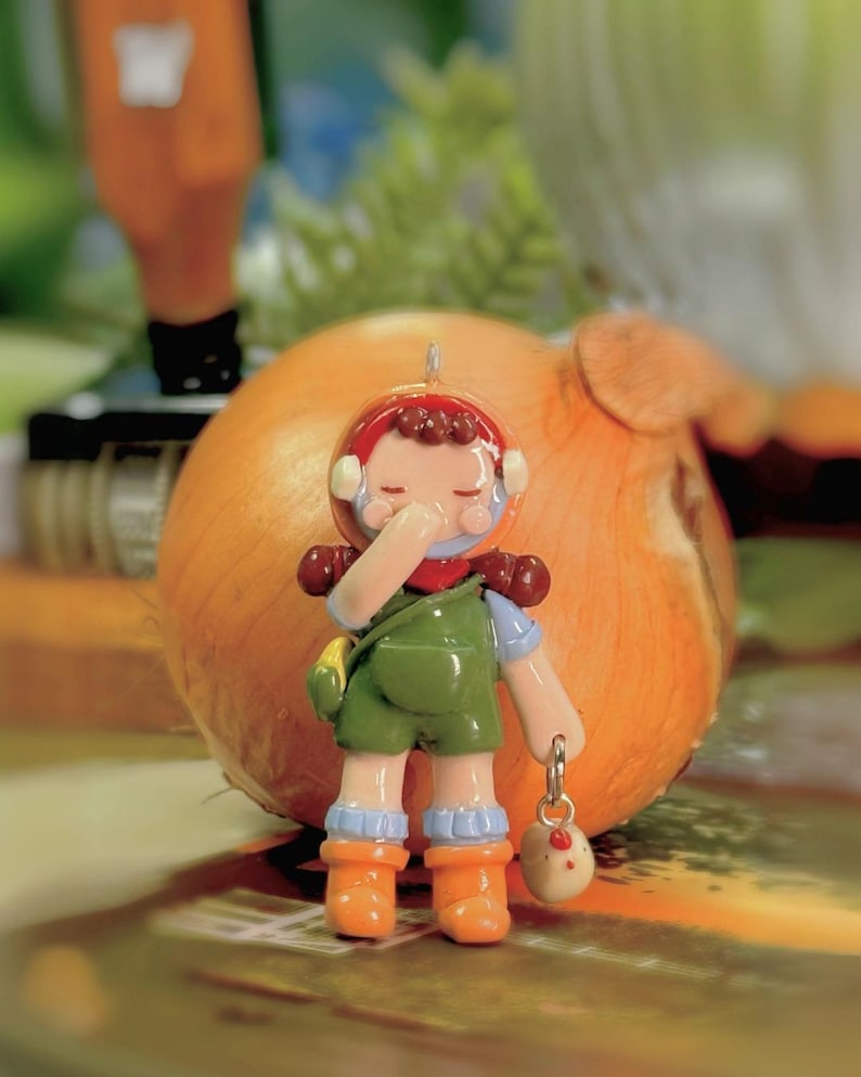 May include: A whimsical figurine of a person in green overalls, orange boots, and a red helmet, holding a small bag and a charm. The charm is a small egg with a chicken design. The figurine is set against a blurred background with an onion.