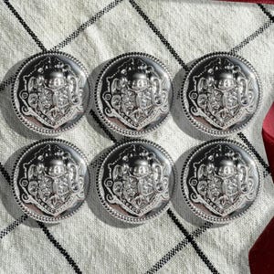 May include: Six silver-toned buttons with an ornate design. Each button features a raised, detailed pattern with a central motif. The buttons are arranged in two rows of three, on a white and black checkered fabric.