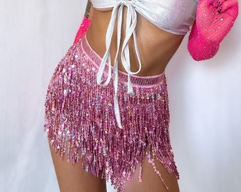 May include: A pink cowboy hat with silver trim and a matching pink bandana. The outfit includes a silver tube top, a pink sequined skirt, and pink gloves. The ensemble is a festival or party outfit.