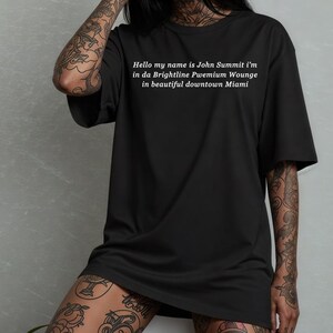 Op de afbeelding: Zwart oversized T-shirt met witte tekst: "Hello my name is John Summit i'm in da Brightline Pwemium Wounge in beautiful downtown Miami." Een casual unisex stijl.