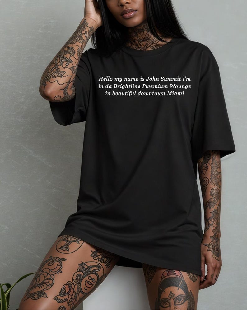 Op de afbeelding: Zwart oversized T-shirt met witte tekst: "Hello my name is John Summit i'm in da Brightline Pwemium Wounge in beautiful downtown Miami." Een casual unisex stijl.