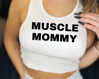 May include: White ribbed vest top featuring the text "MUSCLE MOMMY" in black. The top is worn with light blue denim jeans. The image is a close-up shot.