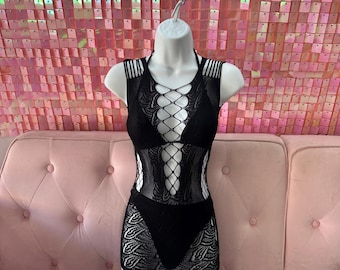 May include: Black lingerie set on a white mannequin. The set includes a halter top with crisscross detailing, a matching skirt, and a thong. The lingerie is made of a combination of lace and mesh fabrics. The background features a pink sequined wall and a pink sofa.
