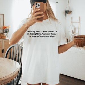 Op de afbeelding: Wit T-shirt met de tekst "Hello my name is John Summit i'm in da Brightline Pwemium Wounge in beautiful downtown Miami." Een zwarte baseball cap met "COWBOY HAT" erop.