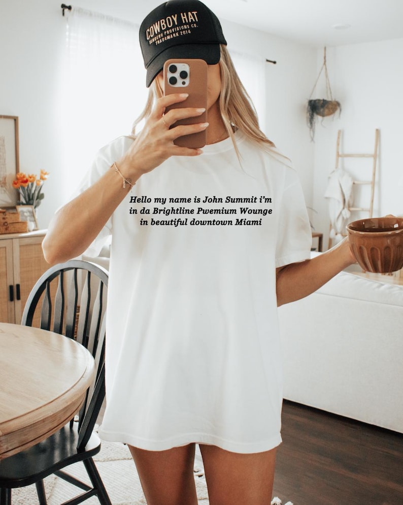 Op de afbeelding: Wit T-shirt met de tekst "Hello my name is John Summit i'm in da Brightline Pwemium Wounge in beautiful downtown Miami." Een zwarte baseball cap met "COWBOY HAT" erop.