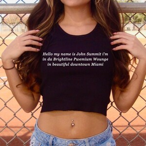 Op de afbeelding: Een zwarte crop top met de tekst "Hello my name is John Summit i'm in da Brightline Pwemium Wounge in beautiful downtown Miami." Gedragen met een blauwe jeans. Een navelpiercing is zichtbaar.