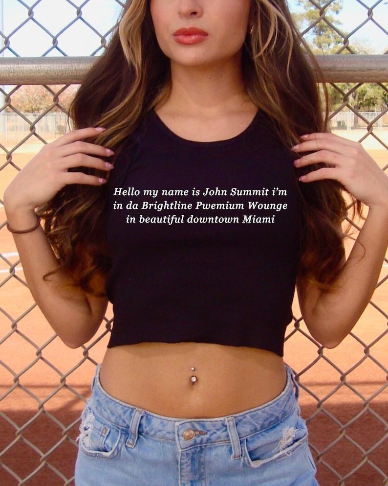 Op de afbeelding: Een zwarte crop top met de tekst "Hello my name is John Summit i'm in da Brightline Pwemium Wounge in beautiful downtown Miami." Gedragen met een blauwe jeans. Een navelpiercing is zichtbaar.