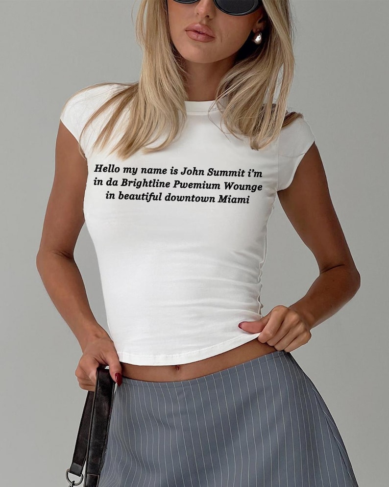 Op de afbeelding: Wit T-shirt met korte mouwen met de tekst "Hello my name is John Summit i'm in da Brightline Pwemium Wounge in beautiful downtown Miami" in zwart. Het shirt wordt gecombineerd met een grijze krijtstreeprok.