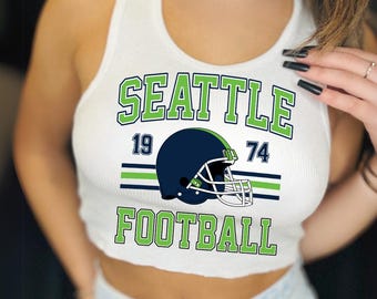 May include: White ribbed vest top with a graphic design. It features the word "SEATTLE" in green block letters, the numbers "19" and "74", and a navy blue football helmet. The word "FOOTBALL" is also in green block letters.