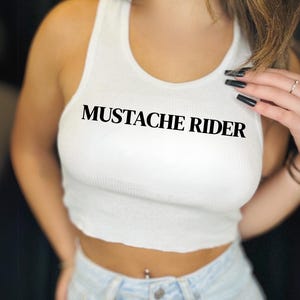 Könnte beinhalten: Weißes geripptes Tanktop mit dem Text "MUSTACHE RIDER" in Schwarz. Das Crop-Top ist ein lässiger Stil, geeignet für warmes Wetter. Es wird mit hellblauen Jeans kombiniert.