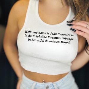 Op de afbeelding: Wit geribbeld tanktop met de tekst "Hello my name is John Summit i'm in da Brightline Pwemium Wounge in beautiful downtown Miami". Gedragen met een lichtblauwe spijkerbroek.