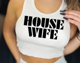 Puede incluir: Top corto blanco acanalado con el texto "HOUSE WIFE" en letras negras en negrita. El top es sin mangas y tiene un escote redondo. La modelo lleva vaqueros azul claro.