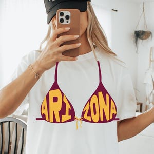 May include: White t-shirt featuring a maroon bikini top design with the word "ARIZONA" in yellow block letters. The shirt is worn with a black baseball cap that says "COWBOY HAT".