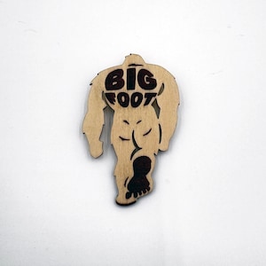 May include: A wooden cutout of Bigfoot walking, with the words "BIG FOOT" printed on the back. The cutout is brown and the text is black.