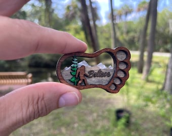 May include: A wooden cutout of a Bigfoot footprint with a scene inside of a Bigfoot walking through a forest with mountains in the background. The text "Believe" is written on the wood.